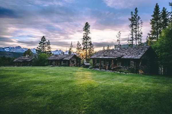 Log cabin lodge and guest rooms on a green lawn with snow-capped mountains in the background at sunset.