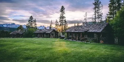 Log cabin lodge and guest rooms on a green lawn with snow-capped mountains in the background at sunset.