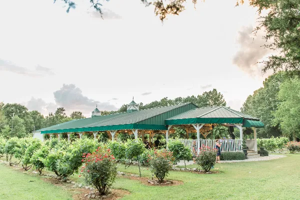 A wide shot of a large event pavilion with a green metal roof and cupolas, situated in a garden with rows of grapevines and flowering shrubs.