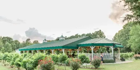 A wide shot of a large event pavilion with a green metal roof and cupolas, situated in a garden with rows of grapevines and flowering shrubs.