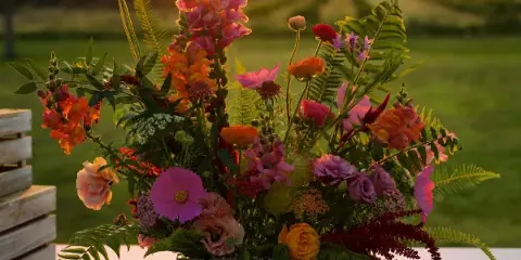 A vibrant floral arrangement with pink snapdragons, orange roses, and ferns in a white rectangular container, set against a blurred background of a green field at sunset.