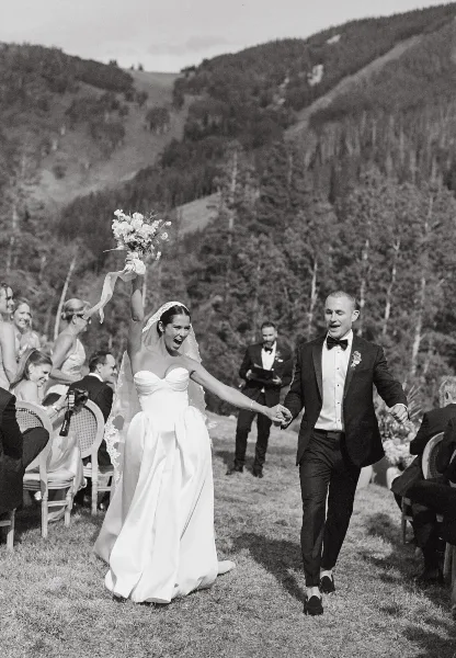 A black and white photograph of a bride and groom walking down the aisle at an outdoor mountain wedding ceremony.