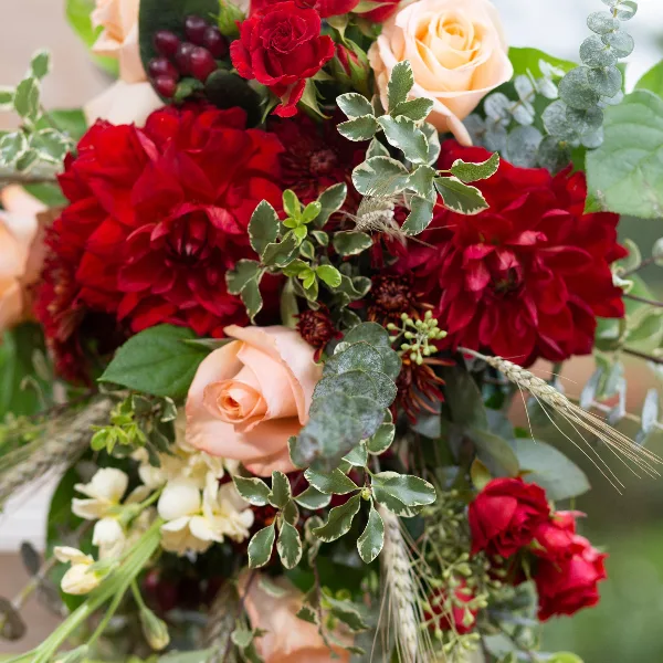 A close-up photograph of a lush floral arrangement featuring deep red dahlias, peach and red roses, and variegated greenery.