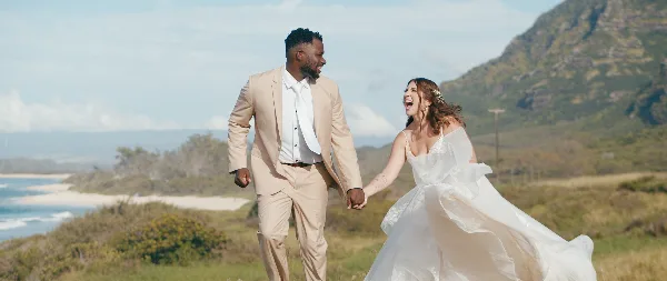 A bride and groom laughing and holding hands while walking outdoors in a scenic, hilly landscape.