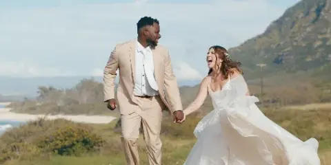 A bride and groom laughing and holding hands while walking outdoors in a scenic, hilly landscape.