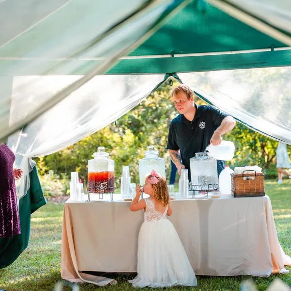 A catering staff member serving drinks from a table with beverage dispensers at an outdoor event.