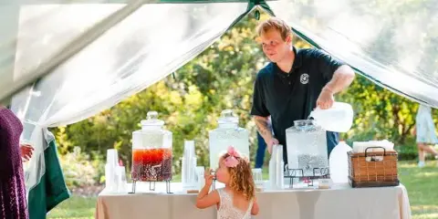 A catering staff member serving drinks from a table with beverage dispensers at an outdoor event.