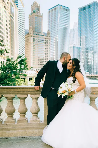 A bride and groom posing together on a balcony with a city skyline in the background.