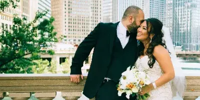 A bride and groom posing together on a balcony with a city skyline in the background.