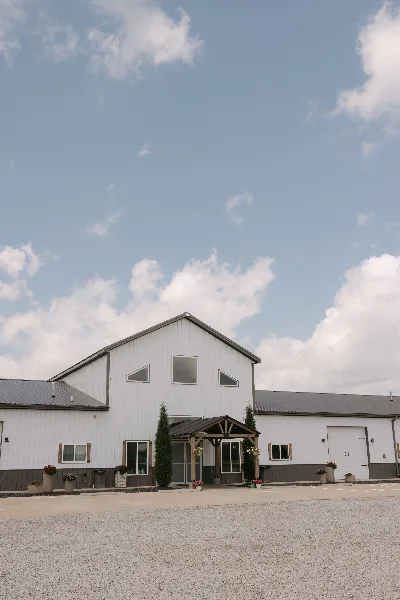 Exterior view of a white barn-style event venue with a covered entrance and gravel parking lot under a blue sky.