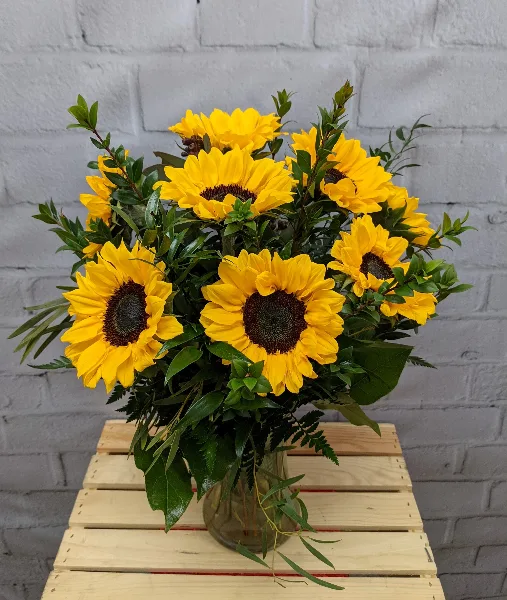 A large bouquet of yellow sunflowers and green foliage arranged in a glass vase on a wooden crate.