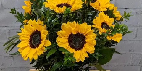 A large bouquet of yellow sunflowers and green foliage arranged in a glass vase on a wooden crate.