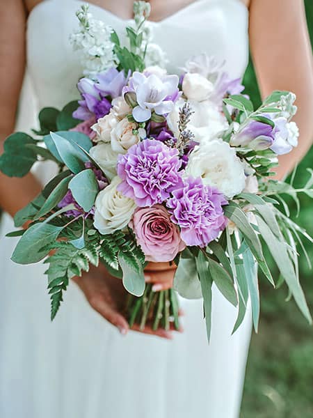 A bride in a white dress holding a hand-tied wedding bouquet featuring purple and white flowers and greenery.