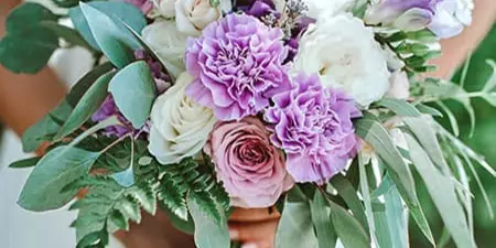 A bride in a white dress holding a hand-tied wedding bouquet featuring purple and white flowers and greenery.
