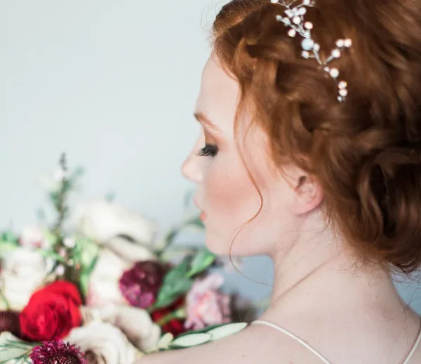 Profile view of a bride with styled red hair, a hair accessory, and makeup, holding a floral bouquet.
