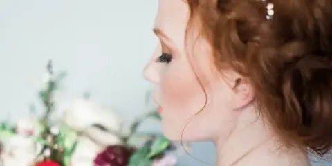 Profile view of a bride with styled red hair, a hair accessory, and makeup, holding a floral bouquet.