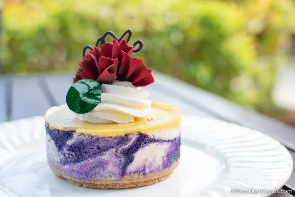 A slice of layered ube cheesecake topped with cream, a green candy, and a red flower garnish.