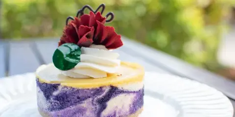 A slice of layered ube cheesecake topped with cream, a green candy, and a red flower garnish.