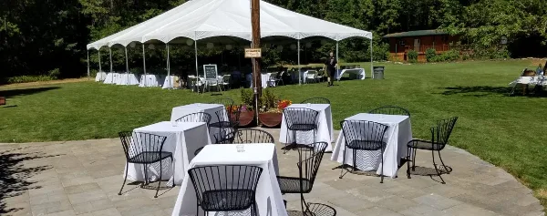 An outdoor wedding venue featuring a large white event tent set up on a green lawn with several tables and chairs arranged for guests.