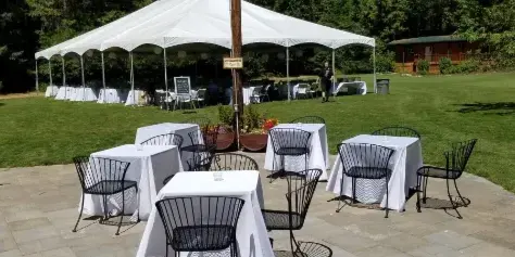An outdoor wedding venue featuring a large white event tent set up on a green lawn with several tables and chairs arranged for guests.