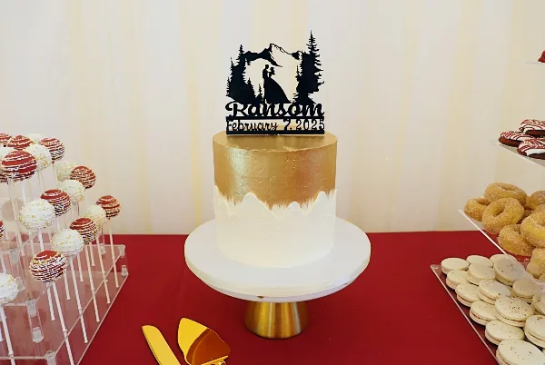 A gold and white wedding cake with a mountain-themed topper displayed on a stand next to a dessert bar with cake pops and macarons.