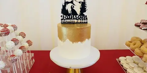 A gold and white wedding cake with a mountain-themed topper displayed on a stand next to a dessert bar with cake pops and macarons.