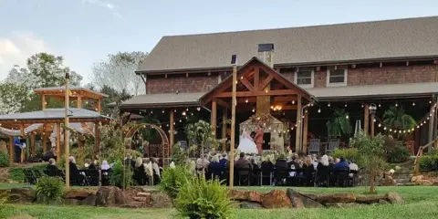 A wedding ceremony taking place on the covered porch of a large wooden venue building with guests seated in the foreground.