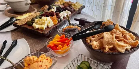 A catering table featuring a large charcuterie board, bowls of vegetable crudités, dips, and fried snacks set on a table with serving utensils.