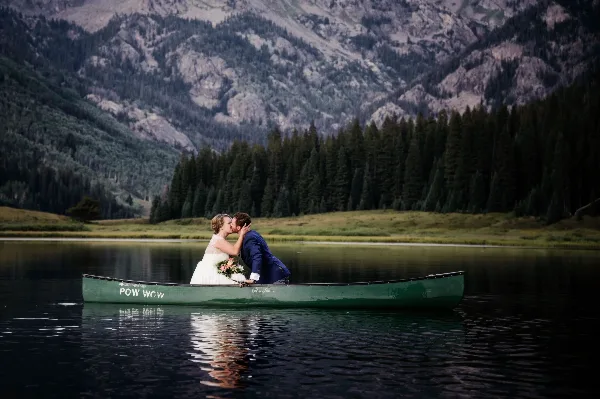 A bride and groom kissing in a green canoe on a calm lake with mountains in the background.