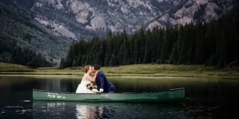 A bride and groom kissing in a green canoe on a calm lake with mountains in the background.
