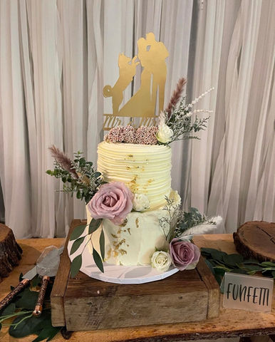 A two-tier white wedding cake decorated with pink and white roses, greenery, and a gold silhouette topper, displayed on a wooden stand.
