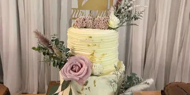 A two-tier white wedding cake decorated with pink and white roses, greenery, and a gold silhouette topper, displayed on a wooden stand.
