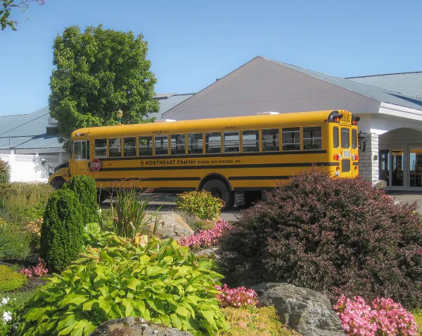 A yellow school bus with Northeast Charter branding parked in front of a white building with a landscaped garden.