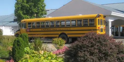 A yellow school bus with Northeast Charter branding parked in front of a white building with a landscaped garden.