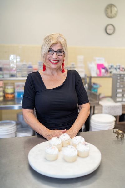 A woman with blonde hair and glasses stands in a bakery kitchen behind a plate of white-frosted cupcakes.