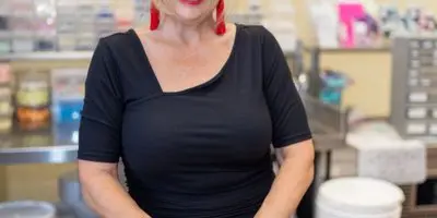 A woman with blonde hair and glasses stands in a bakery kitchen behind a plate of white-frosted cupcakes.