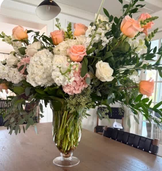 A large, elegant floral arrangement with peach roses, white hydrangeas, and greenery in a clear glass vase on a wooden table.