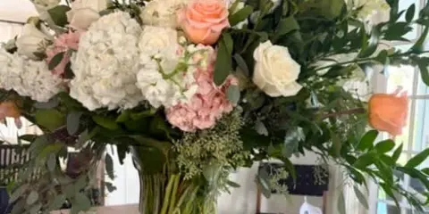 A large, elegant floral arrangement with peach roses, white hydrangeas, and greenery in a clear glass vase on a wooden table.