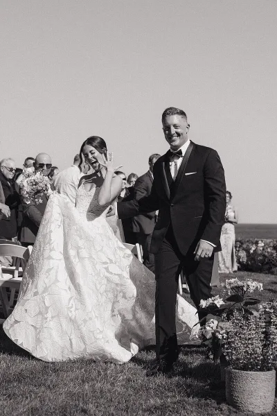 A black and white photo of a bride and groom laughing and walking down an outdoor aisle, with the bride's dress flowing behind her.