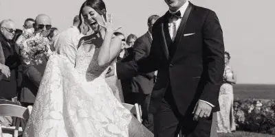 A black and white photo of a bride and groom laughing and walking down an outdoor aisle, with the bride's dress flowing behind her.