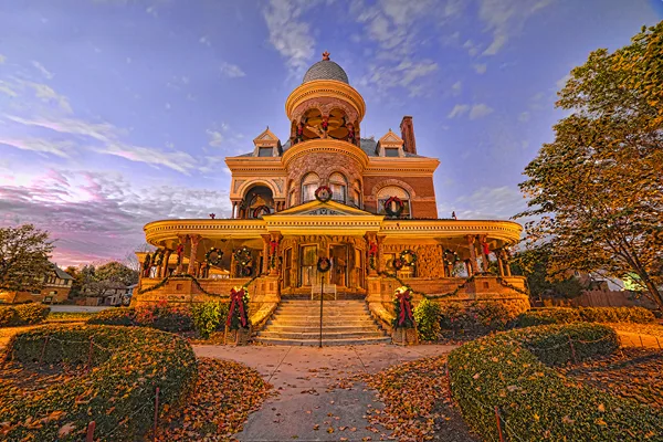 Exterior view of the Seiberling Mansion, a historic Victorian-style building with a prominent dome and wraparound porch, decorated with wreaths and illuminated at dusk.