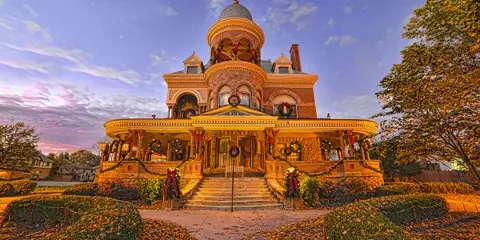 Exterior view of the Seiberling Mansion, a historic Victorian-style building with a prominent dome and wraparound porch, decorated with wreaths and illuminated at dusk.