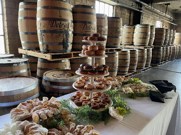 A catering table set up in a warehouse with wooden barrels, featuring a tiered stand of donuts and pastries alongside a platter of powdered pastries.