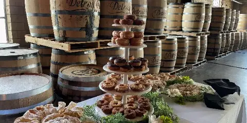 A catering table set up in a warehouse with wooden barrels, featuring a tiered stand of donuts and pastries alongside a platter of powdered pastries.