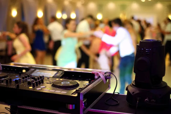 A professional DJ mixer and stage light in the foreground with a blurred crowd dancing in the background.
