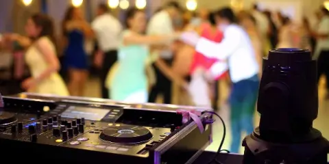 A professional DJ mixer and stage light in the foreground with a blurred crowd dancing in the background.