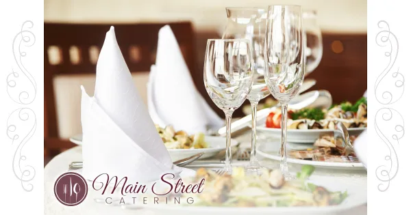 A formal dining table setting featuring plated food, wine glasses, and folded white napkins with the Main Street Catering logo.