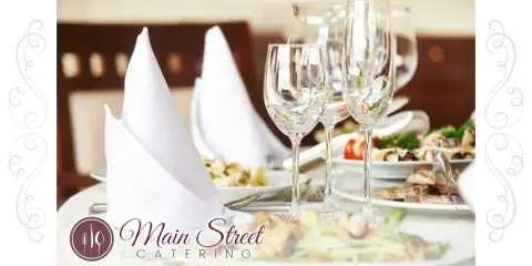 A formal dining table setting featuring plated food, wine glasses, and folded white napkins with the Main Street Catering logo.