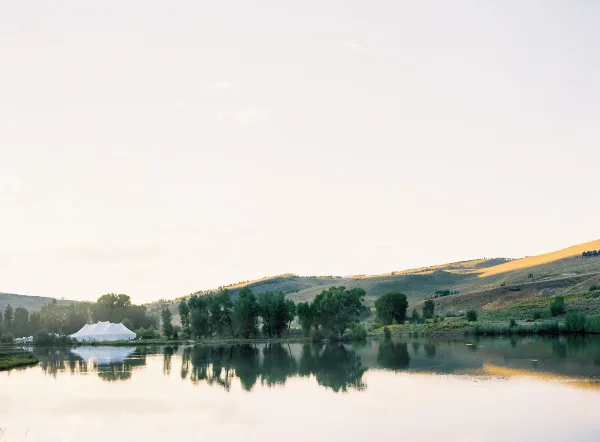 White event tent set up near a lake and rolling hills at an outdoor venue.