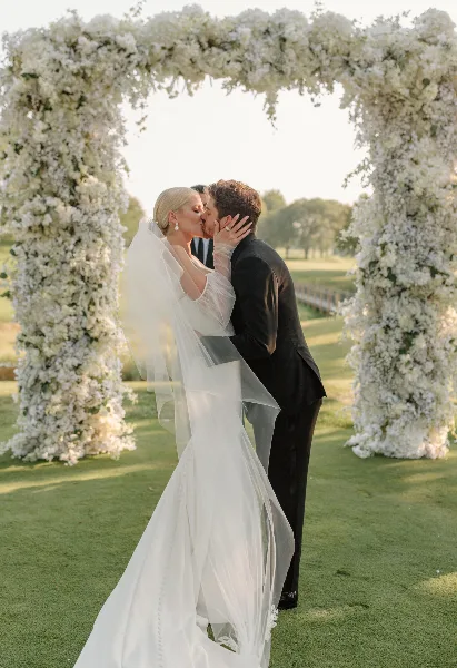 A bride and groom sharing a kiss under a white floral arch at an outdoor wedding venue.